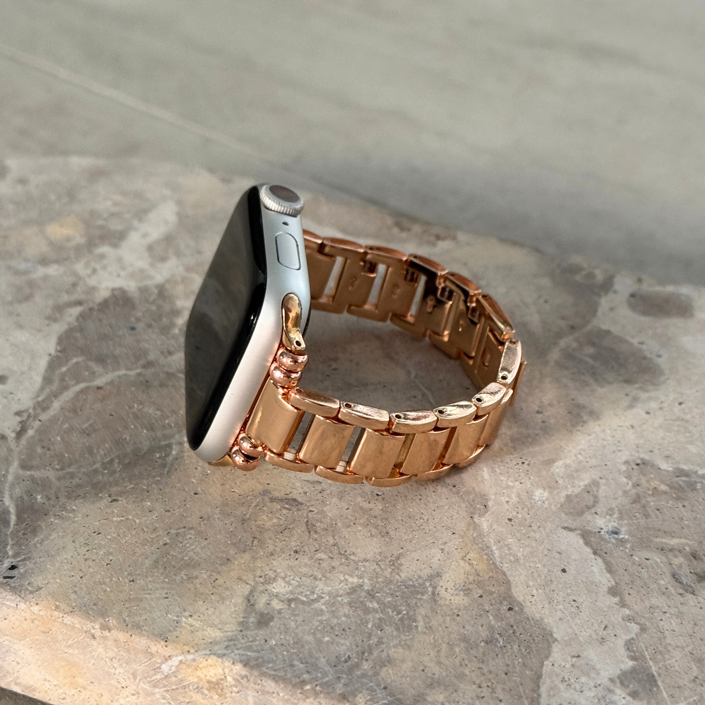 Rose gold slim link Apple Watch band propped upright on a stone surface. Lightweight metal iWatch armband with a polished finish, compatible with Apple Watch 38mm to 49mm, Series 1-11, SE and Ultra