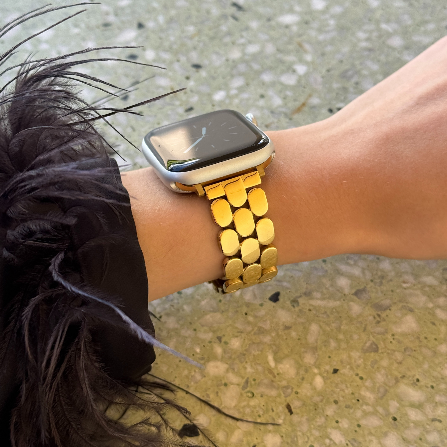 Gold pebble link Apple Watch band worn with a feather trimmed cuff outdoors. Timeless stainless steel iWatch strap with a high shine finish, compatible with Apple Watch 38mm to 49mm, Series 1-11, SE and Ultra