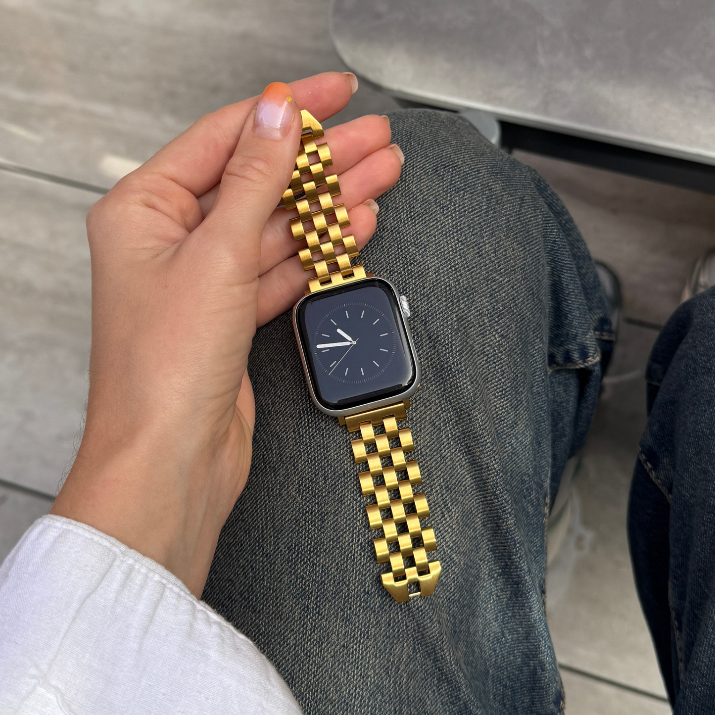 Gold multi link Apple Watch band held in a hand above denim. Unisex stainless steel iWatch armband with a seamless link profile, compatible with Apple Watch 38mm to 49mm, Series 1-11, SE and Ultra