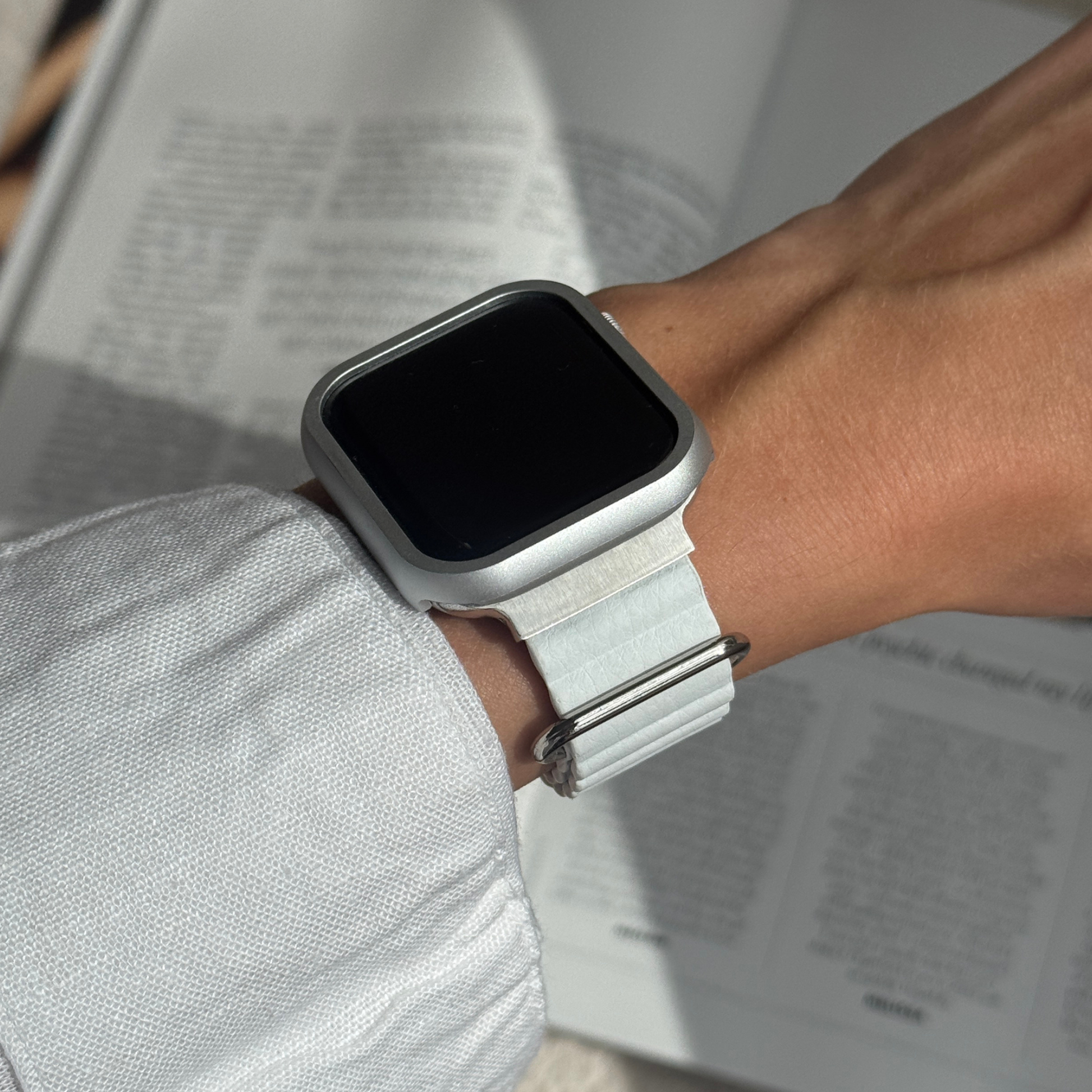 Silver stainless steel Apple Watch case cover worn on a wrist above an open book. Durable protective bumper with a refined finish, available for Apple Watch 38mm to 49mm, Series 1-11, SE and Ultra