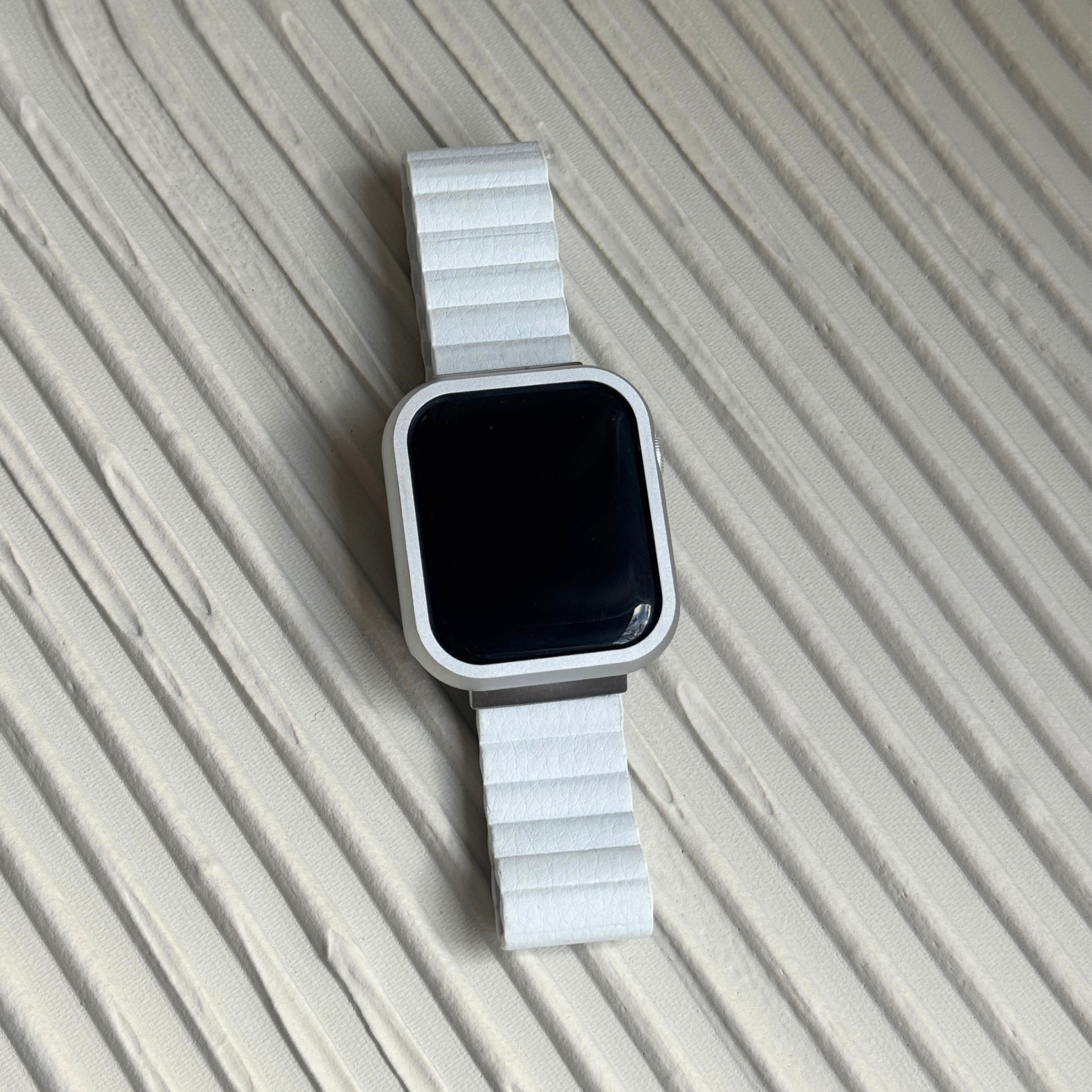 Silver stainless steel Apple Watch case cover shown on a ribbed beige surface with a white band. Refined protective bumper, available for Apple Watch 38mm to 49mm, Series 1-11, SE and Ultra