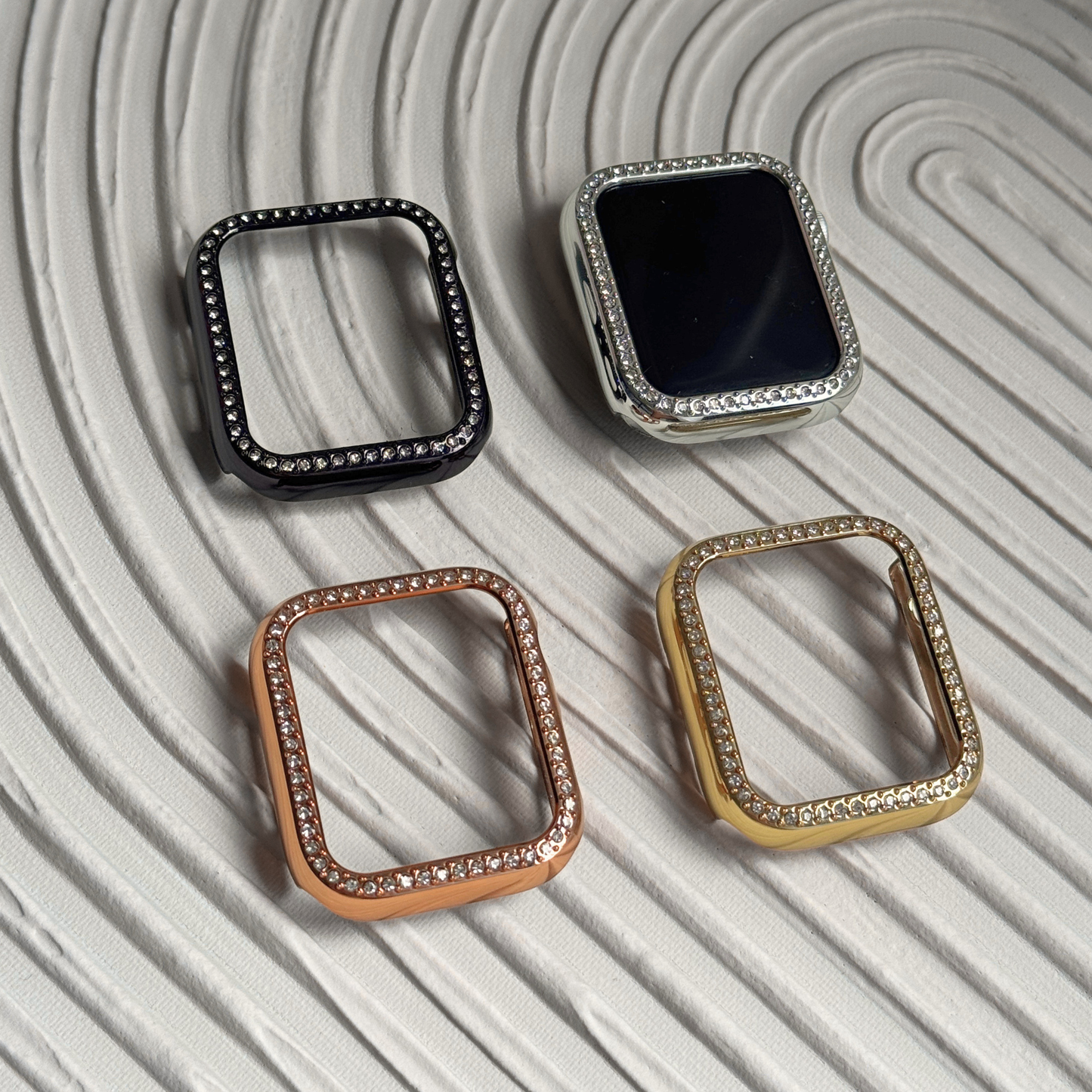 Four square-shaped watch cases in black, silver, rose gold, and gold on a textured light surface.