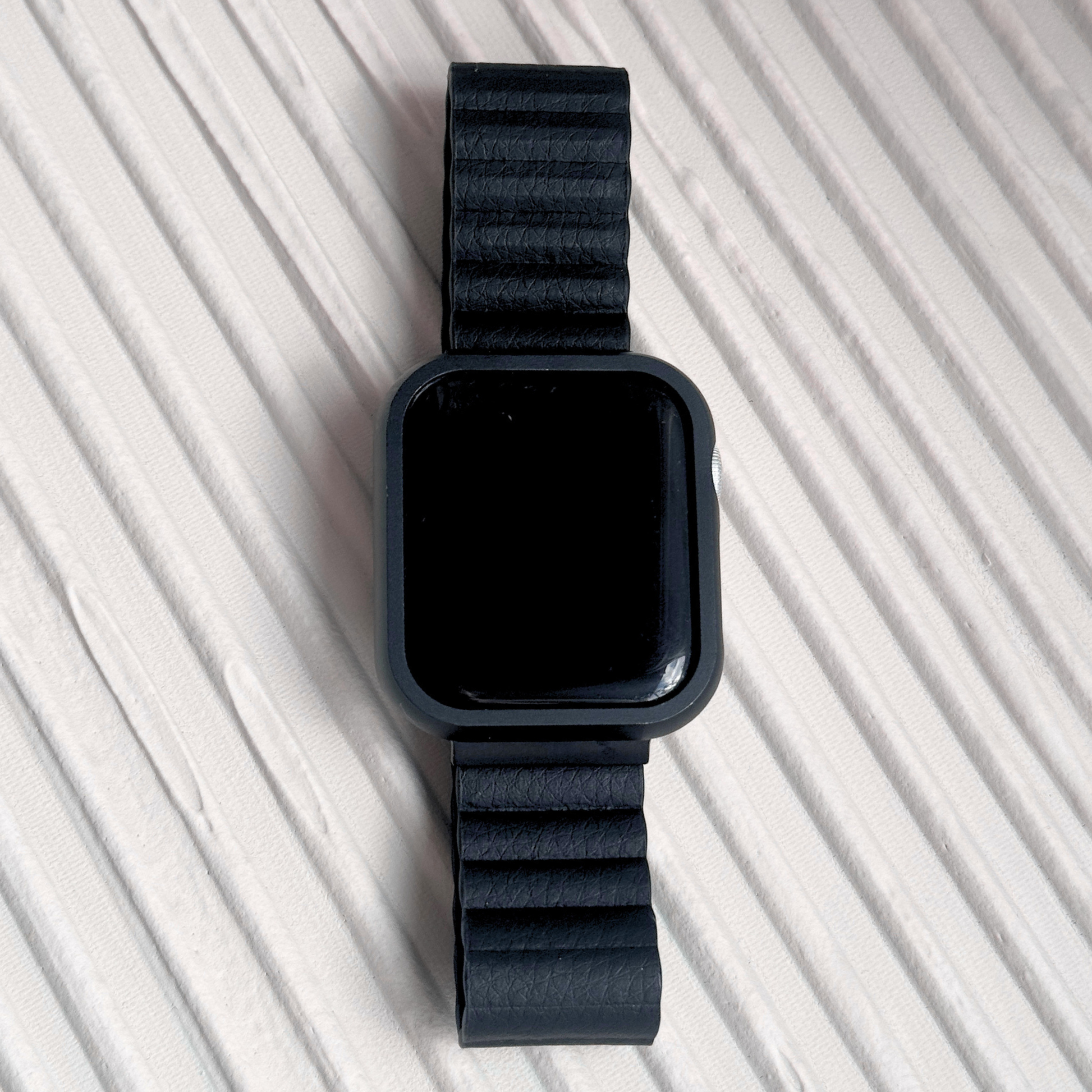 Black stainless steel Apple Watch case cover shown on a ribbed white surface. Premium protective bumper with a refined finish, available for Apple Watch 38mm to 49mm, Series 1-11, SE and Ultra