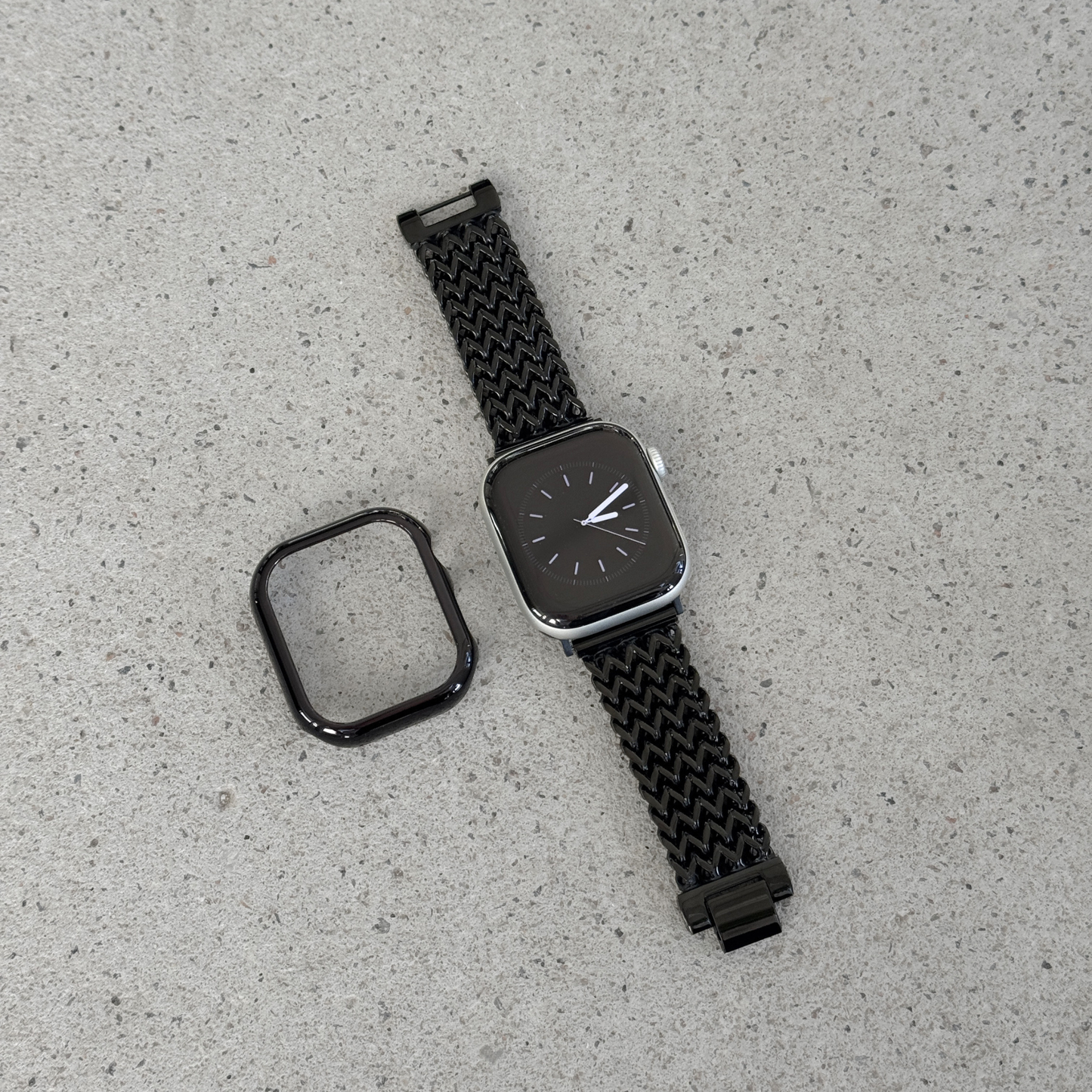 A black smartwatch with a woven black mesh strap and a separate black protective case, displayed on a light textured surface.