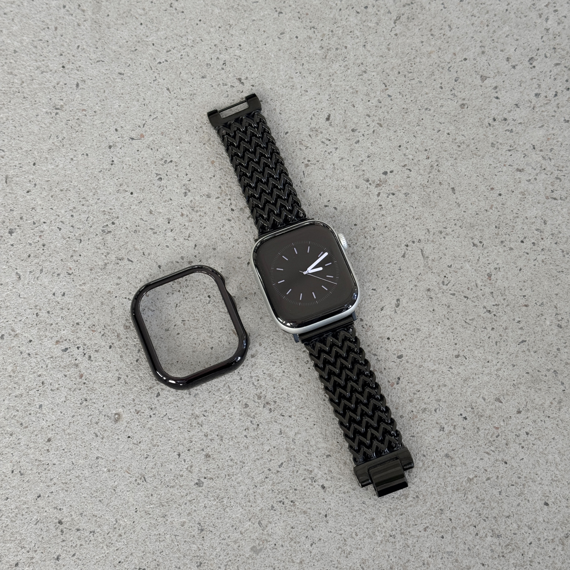 A black smartwatch with a woven black mesh strap and a separate black protective case, displayed on a light textured surface.