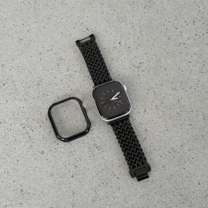 A black smartwatch with a woven black mesh strap and a separate black protective case, displayed on a light textured surface.