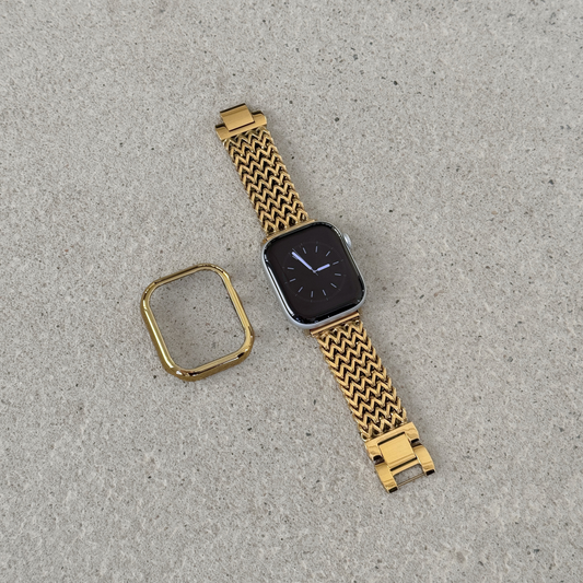 A gold tone metal mesh Apple watch band with a matching gold bezel protector, shown on an apple smartwatch against a textured concrete background.