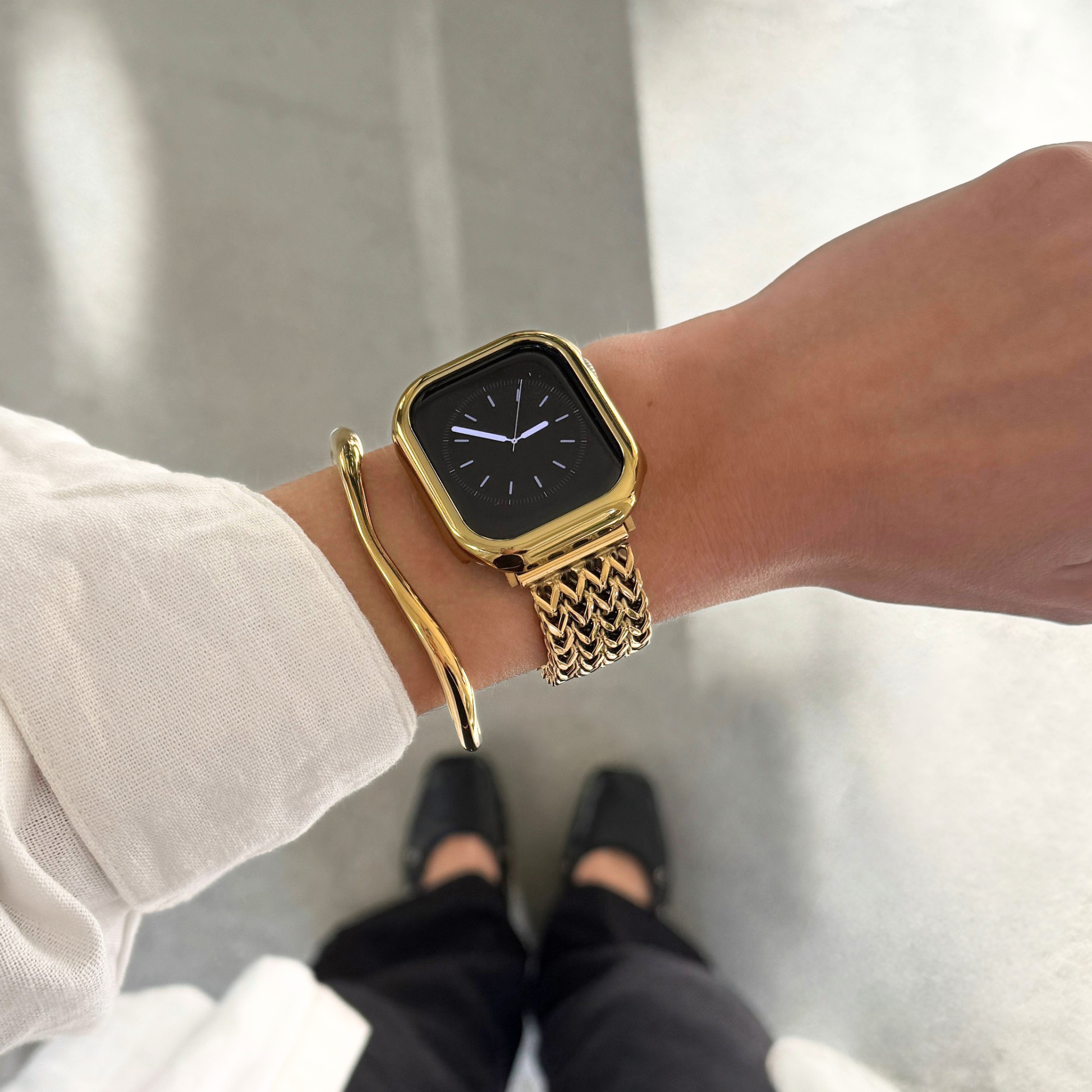A gold toned textured Apple Watch mesh band, bundled with a matching gold bezel case and cuff bracelet, worn on a person's wrist. The person is also wearing a long-sleeve top, with dark pants and shoes visible below against a light  floor.