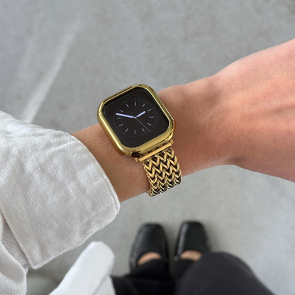 A gold smartwatch with a gold mesh band worn on a person's wrist, shown in a lifestyle setting with concrete floor and stylish shoes visible.