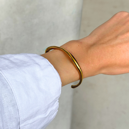 A polished gold toned metal cuff bracelet worn on a person's wrist, shown against a neutral light background.