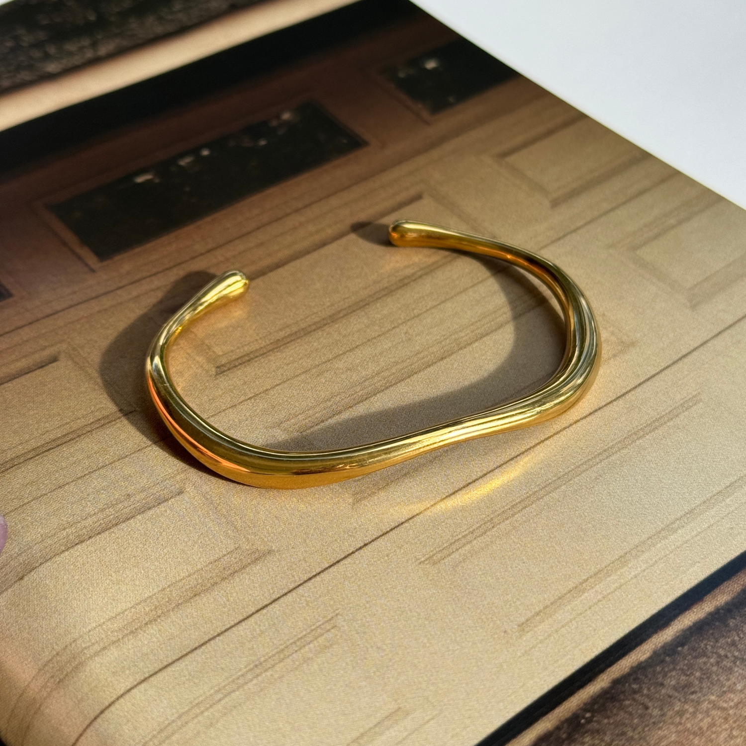 A polished gold toned open cuff bangle bracelet with a wavy, organic curve, resting on a textured beige surface.