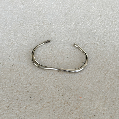 A polished silver toned metal cuff with a wavy, organic curve, displayed on a textured surface.