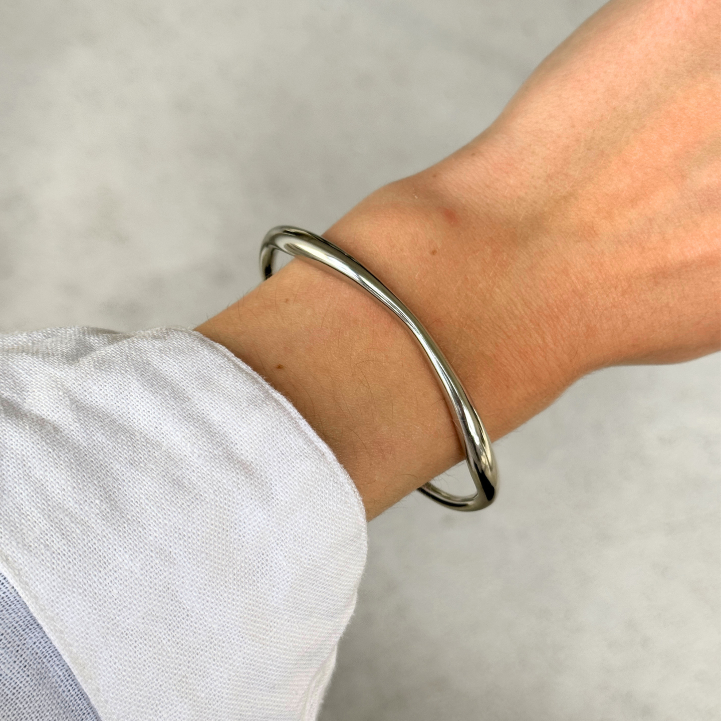 A polished silver toned metal bangle bracelet worn on a person's wrist, shown against a light background with a linen shirt cuff visible.