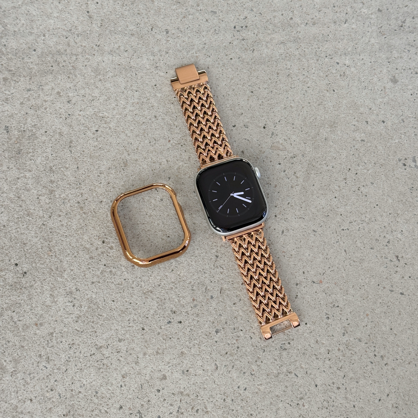 A rose gold metal mesh Apple watch band with a matching square bezel, shown on a smartwatch alongside its detached strap end, placed on a textured concrete surface.