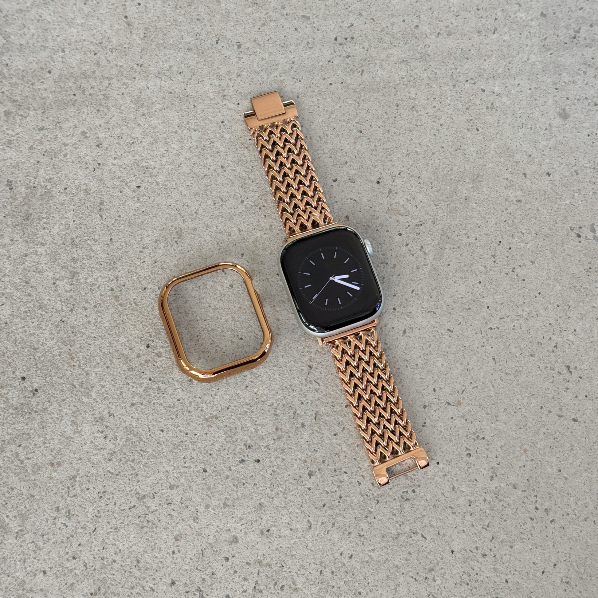 A rose gold metal mesh Apple watch band with a matching square bezel, shown on a smartwatch alongside its detached strap end, placed on a textured concrete surface.