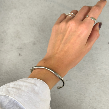 A silver toned metal hinged cuff bracelet worn on a person's wrist, with their hand resting against a light textured background.