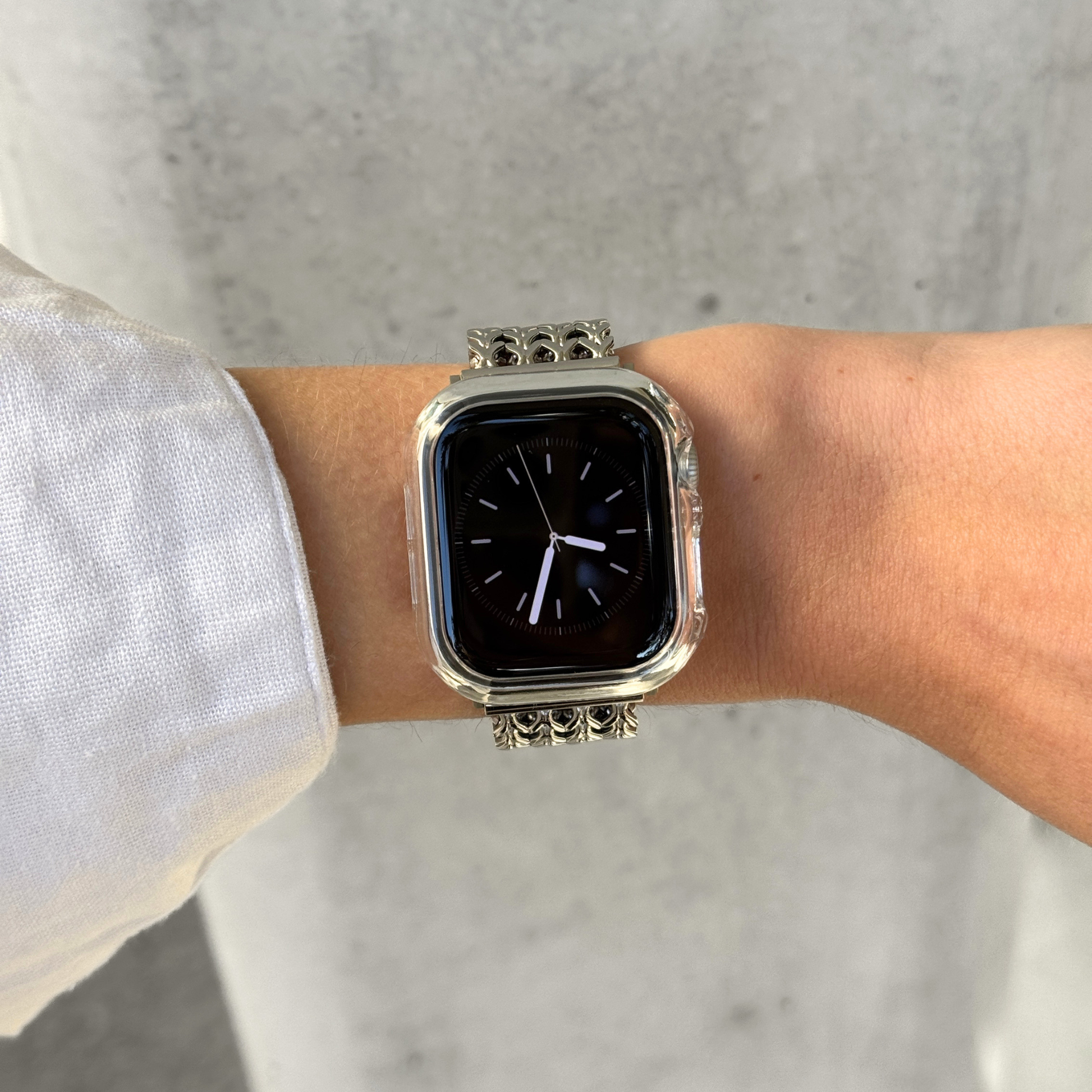 An Apple Watch with a clear bumper case with a decorative chain strap worn on a person's wrist, shown against a light concrete wall.