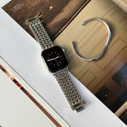 A silver mesh metal watch band attached to an Apple smartwatch with a minimal display, styled on top of an open book next to a silver cuff bracelet.