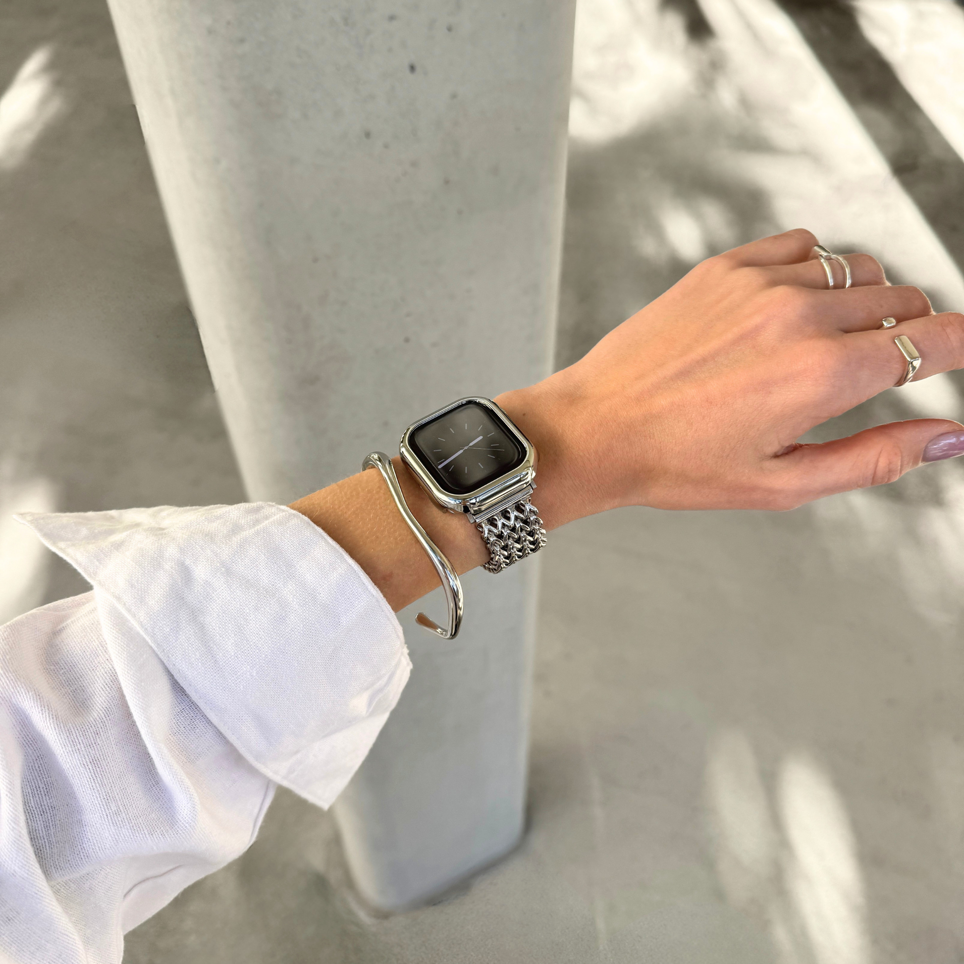 A silver metal Apple watch link strap with a matching silver-toned cuff bracelet and a silver case cover, worn on a person's wrist, styled with a silver bangle and rings, set against a concrete pillar background.