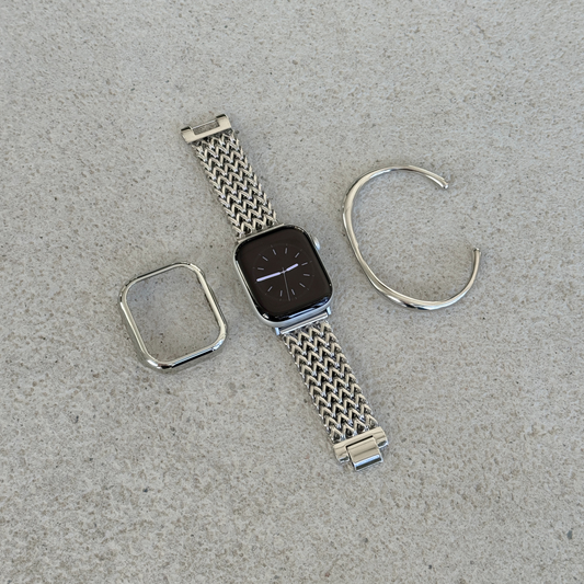 A silver stainless steel mesh Apple watch band with a matching silver bezel protector and a matching cuff bracelet, arranged on a textured surface next to an Apple Watch.