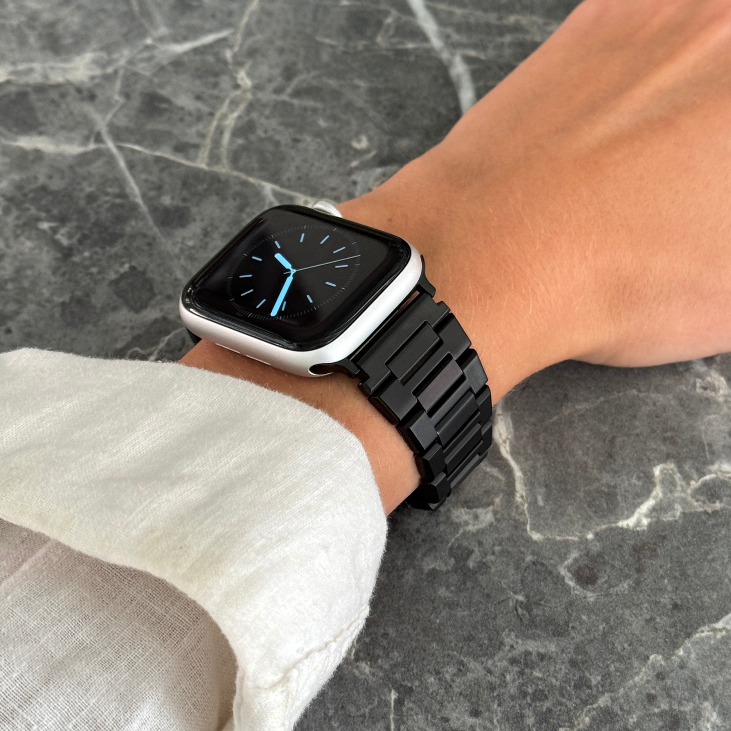 Black link Apple Watch band worn on a wrist above dark marble. Modern unisex stainless steel iWatch bracelet, compatible with Apple Watch 38mm to 49mm, Series 1-11, SE and Ultra