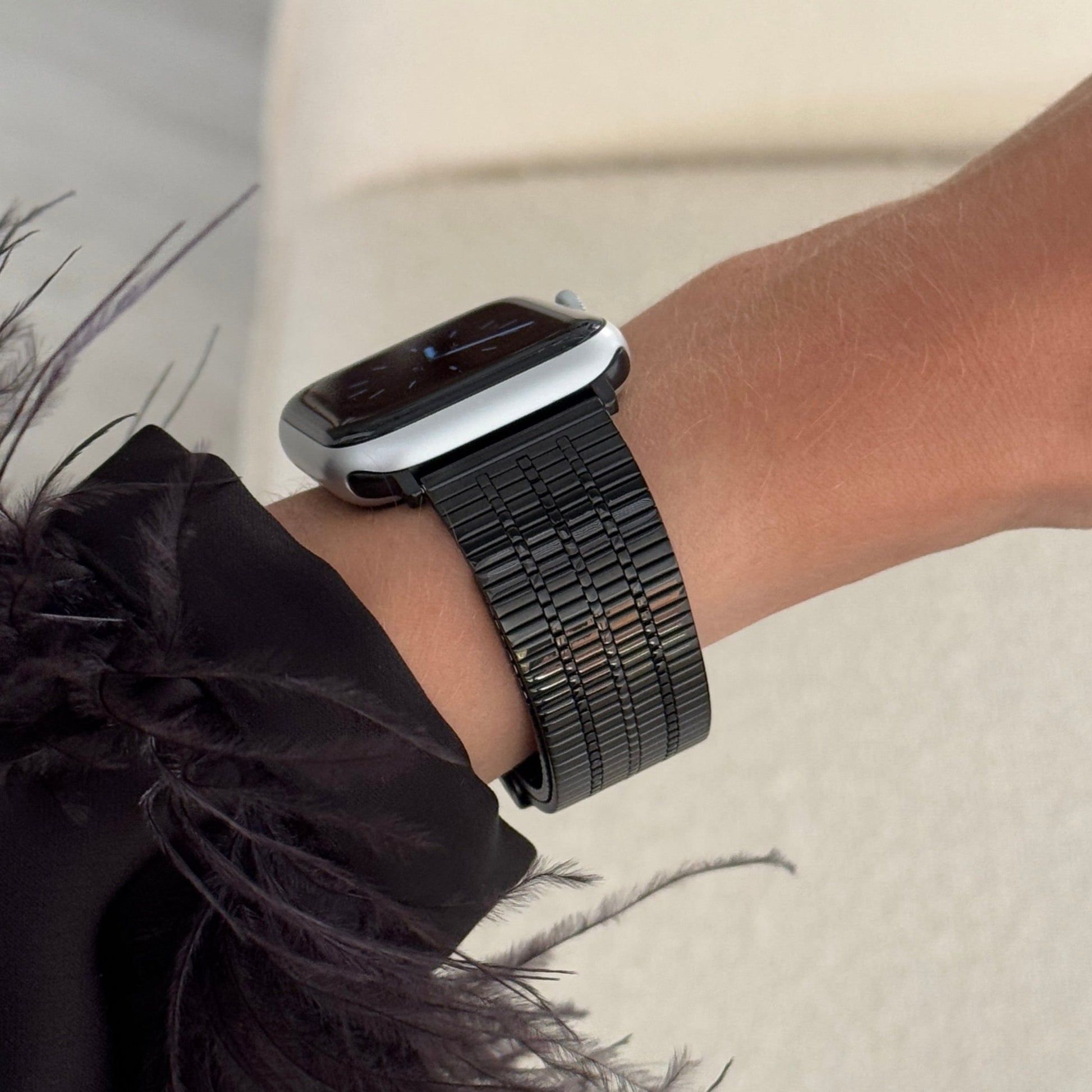Black flat woven Apple Watch band worn with a feather trimmed cuff on a cream sofa. Unisex stainless steel iWatch bracelet, compatible with Apple Watch 38mm to 49mm, Series 1-11, SE and Ultra