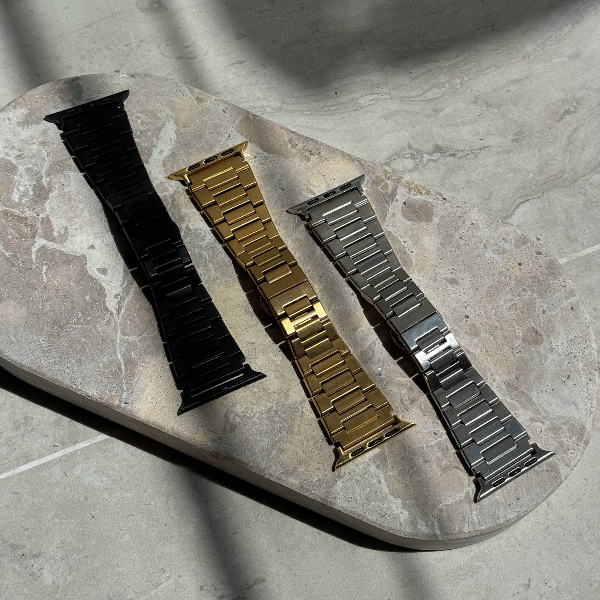 Black, gold and silver link Apple Watch bands laid out on a stone tray. Waterproof stainless steel iWatch straps, compatible with Apple Watch 38mm to 49mm, Series 1-11, SE and Ultra