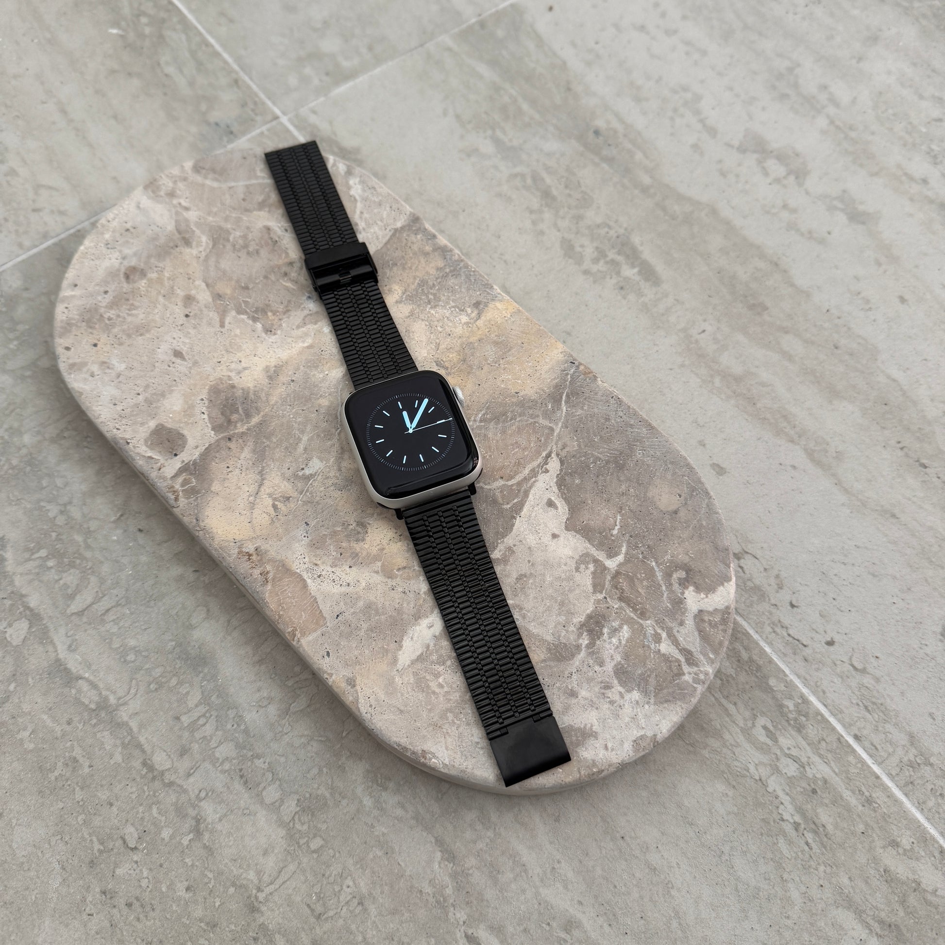 Black flat woven Apple Watch band lying flat on a stone tray. Lightweight stainless steel iWatch strap, compatible with Apple Watch 38mm to 49mm, Series 1-11, SE and Ultra