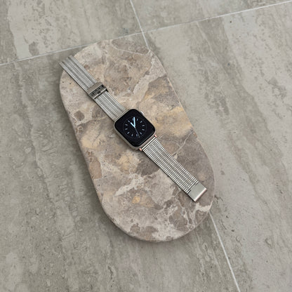 Silver flat woven Apple Watch band lying flat on a stone tray. Stainless steel iWatch bracelet with an easy to use clasp, compatible with Apple Watch 38mm to 49mm, Series 1-11, SE and Ultra
