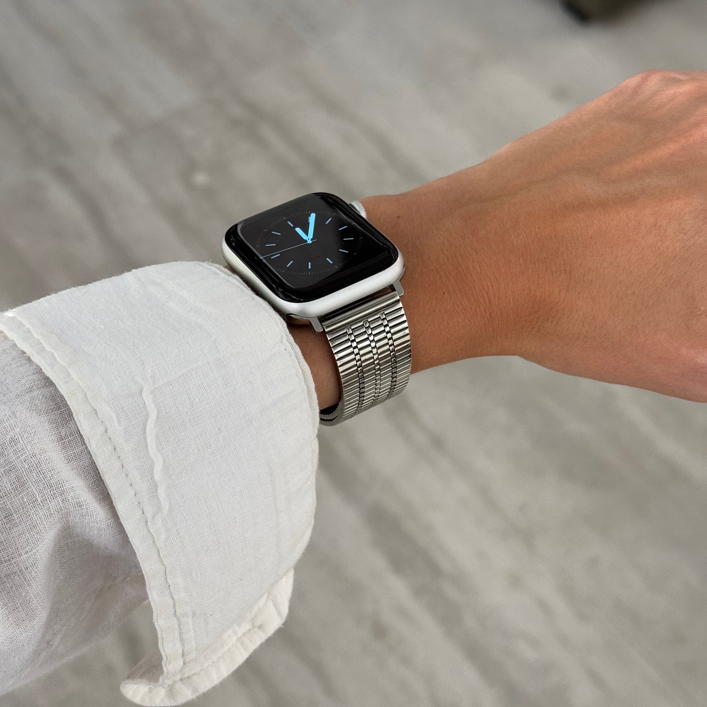 Silver flat woven Apple Watch band worn on a wrist with a white linen sleeve. Stainless steel iWatch bracelet with an easy to use clasp, compatible with Apple Watch 38mm to 49mm, Series 1-11, SE and Ultra