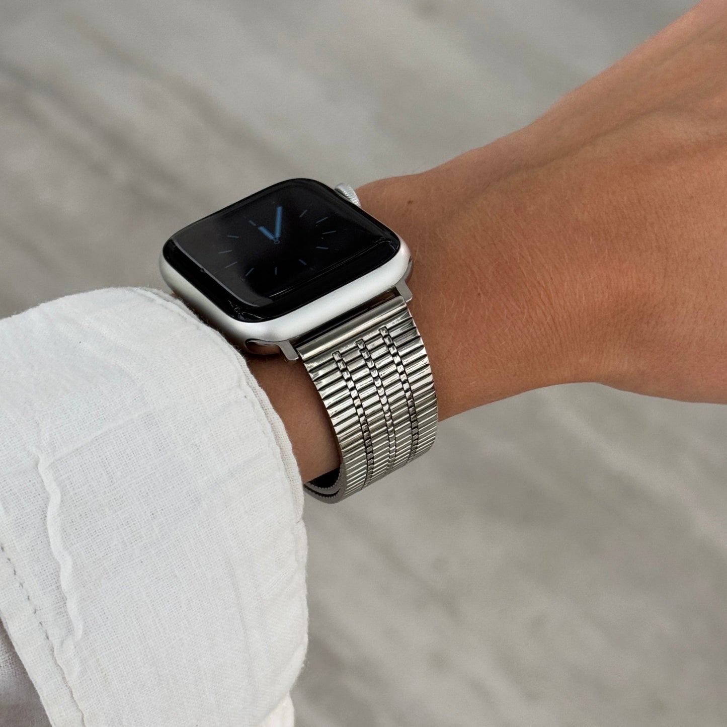 Silver flat woven Apple Watch band worn on a wrist against a stone background. Waterproof stainless steel iWatch strap, compatible with Apple Watch 38mm to 49mm, Series 1-11, SE and Ultra