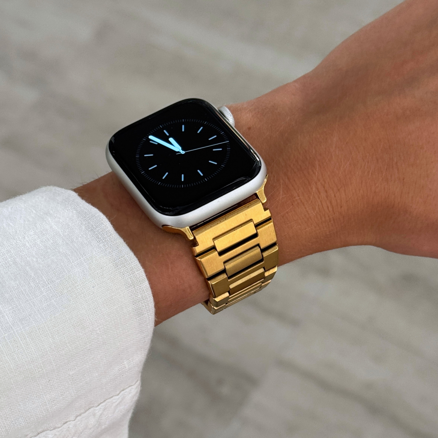 Gold link Apple Watch band worn on a wrist with a white linen cuff. Sleek stainless steel iWatch strap with a brushed metal finish, compatible with Apple Watch 38mm to 49mm, Series 1-11, SE and Ultra