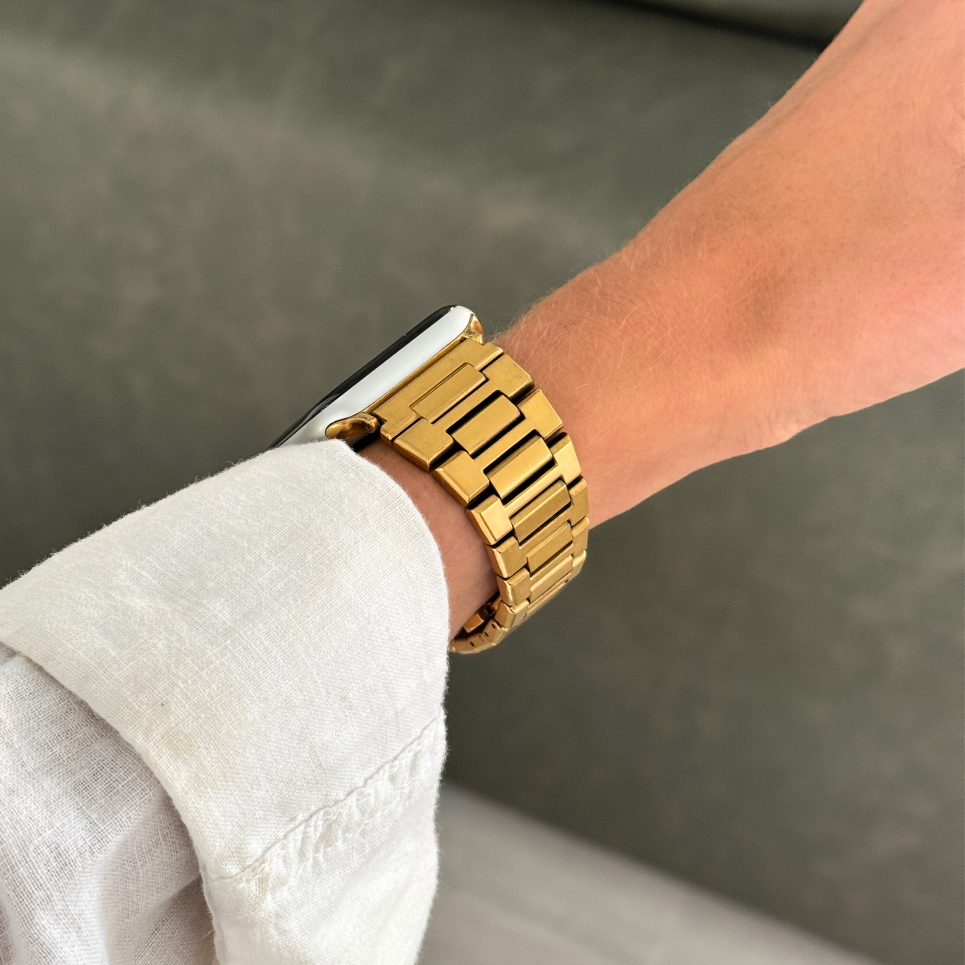 Gold link Apple Watch band worn with a white linen sleeve beside a green sofa. Modern stainless steel iWatch bracelet, compatible with Apple Watch 38mm to 49mm, Series 1-11, SE and Ultra
