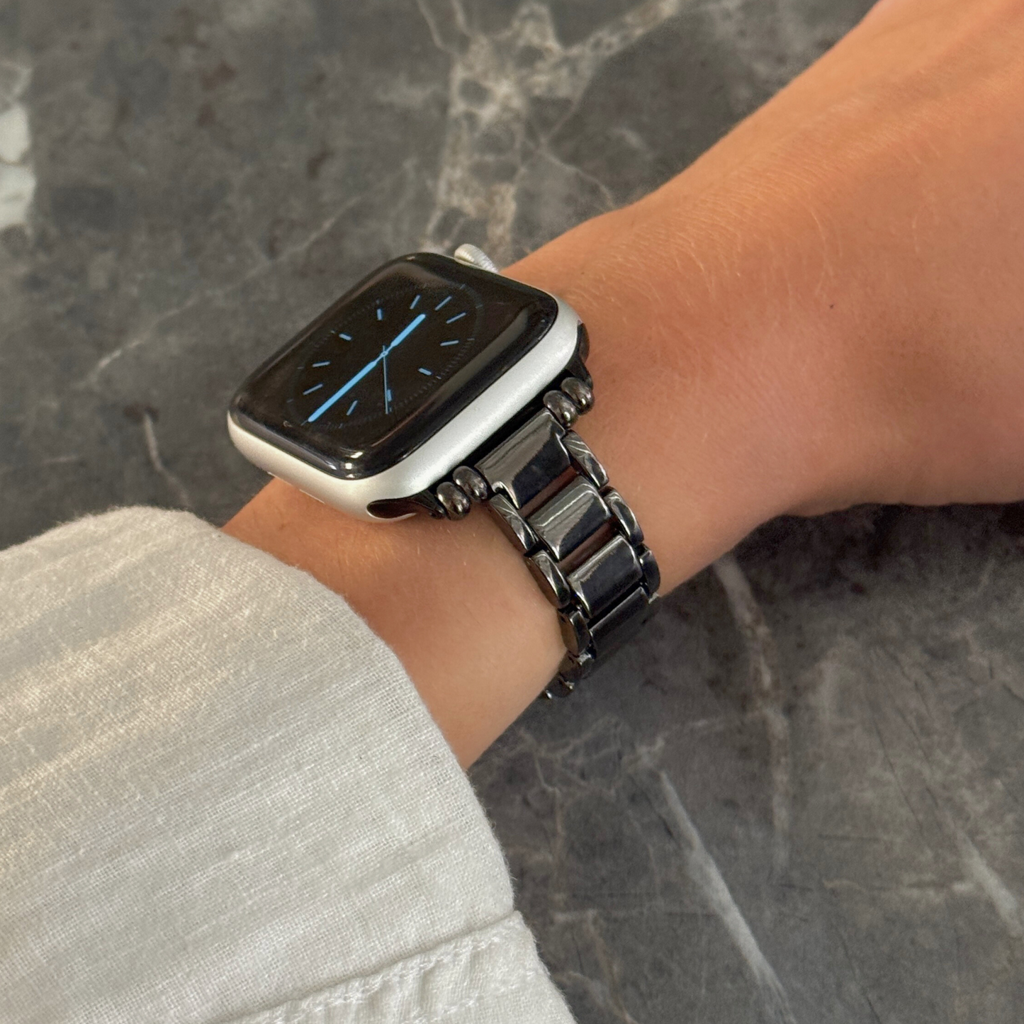 Black slim link Apple Watch band worn on a wrist above grey marble. Sleek feminine metal iWatch strap, compatible with Apple Watch 38mm to 49mm, Series 1-11, SE and Ultra