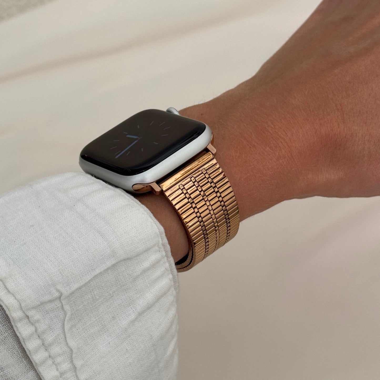 Rose gold flat woven Apple Watch band worn on a wrist with a white linen cuff. Lightweight stainless steel iWatch bracelet, compatible with Apple Watch 38mm to 49mm, Series 1-11, SE and Ultra