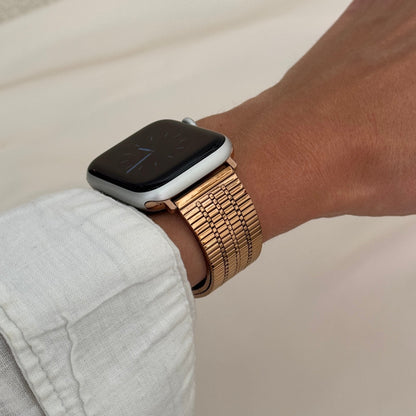 Rose gold flat woven Apple Watch band worn on a wrist with a white linen cuff. Lightweight stainless steel iWatch bracelet, compatible with Apple Watch 38mm to 49mm, Series 1-11, SE and Ultra