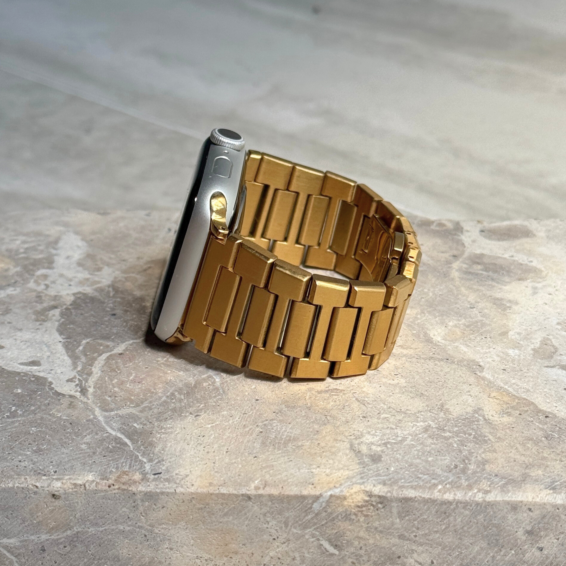 Gold link Apple Watch band propped upright on a stone surface. Waterproof stainless steel iWatch bracelet with a brushed finish, compatible with Apple Watch 38mm to 49mm, Series 1-11, SE and Ultra