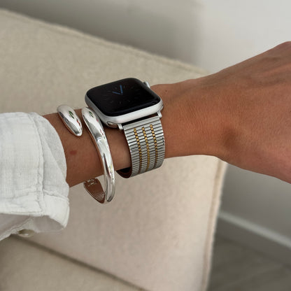 Silver gold flat woven Apple Watch band worn with chunky cuff bangles on a cream sofa. Stainless steel iWatch strap for effortless wear, compatible with Apple Watch 38mm to 49mm, Series 1-11, SE and Ultra