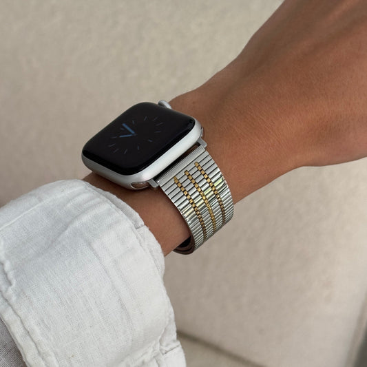 Silver gold flat woven Apple Watch band worn on a wrist with a white linen cuff. Unisex stainless steel iWatch bracelet, compatible with Apple Watch 38mm, 40mm, 41mm, 42mm, 44mm, 45mm, 46mm and 49mm, Series 1-11, SE and Ultra