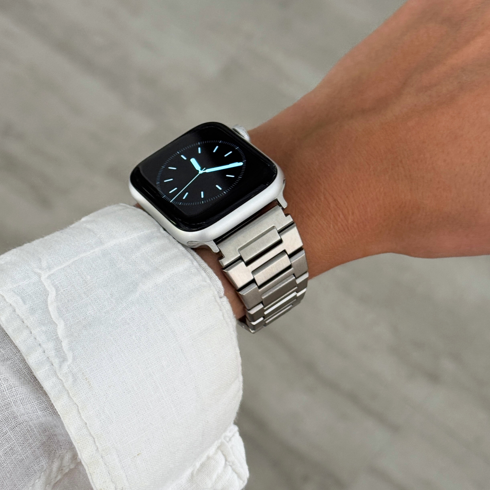 Silver link Apple Watch band worn on a wrist with a white linen cuff. Unisex stainless steel iWatch strap with a brushed metal finish, compatible with Apple Watch 38mm to 49mm, Series 1-11, SE and Ultra