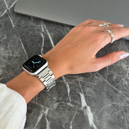 Silver link Apple Watch band worn on a wrist with rings above dark marble. Durable stainless steel iWatch bracelet, compatible with Apple Watch 38mm to 49mm, Series 1-11, SE and Ultra