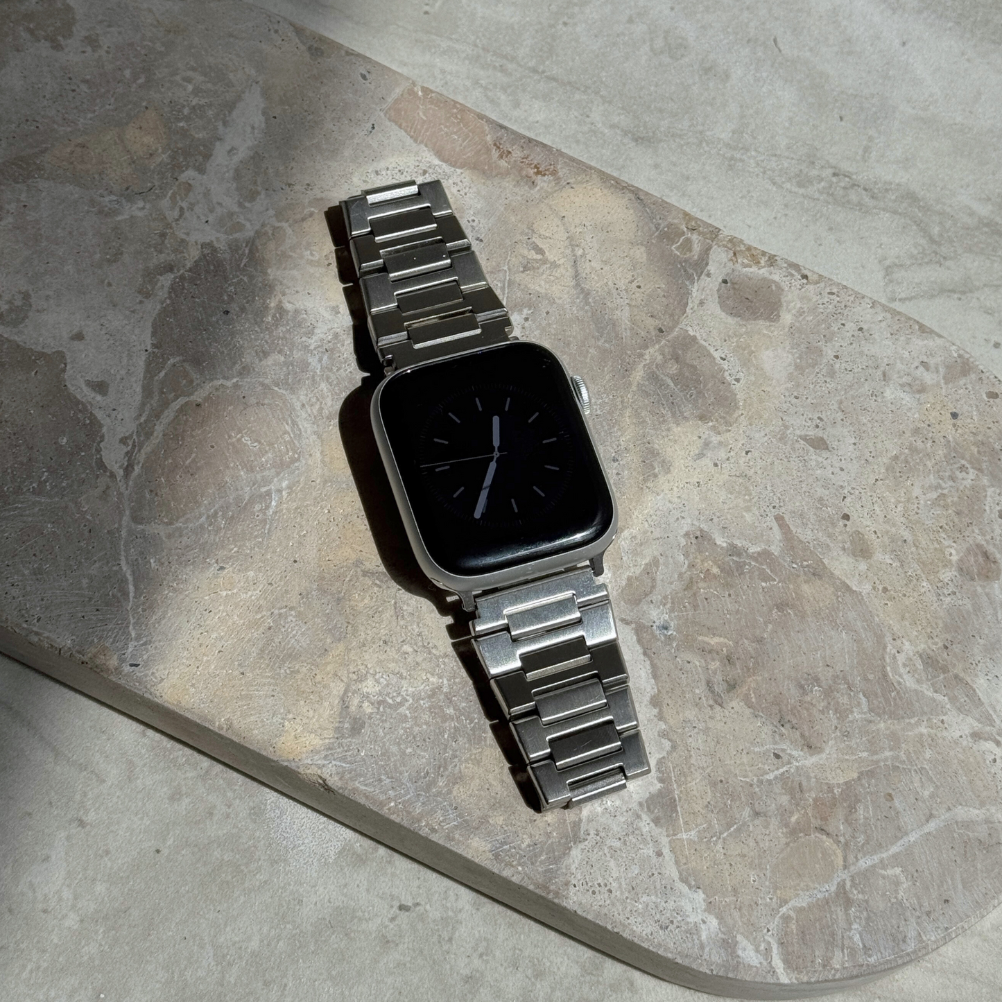 Silver link Apple Watch band lying flat on a stone tray. Waterproof stainless steel iWatch bracelet, durable for everyday wear, compatible with Apple Watch 38mm to 49mm, Series 1-11, SE and Ultra