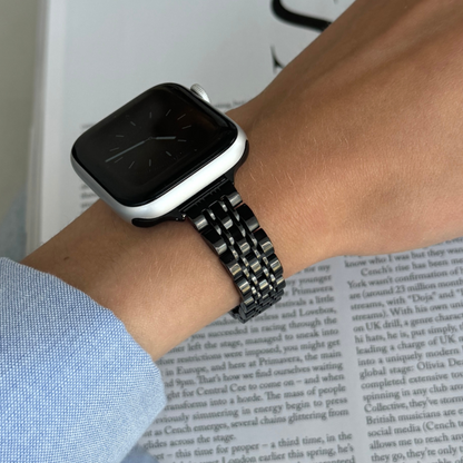 Black woven link Apple Watch band worn on a wrist above an open newspaper, stainless steel bracelet for Apple Watch 38mm to 49mm