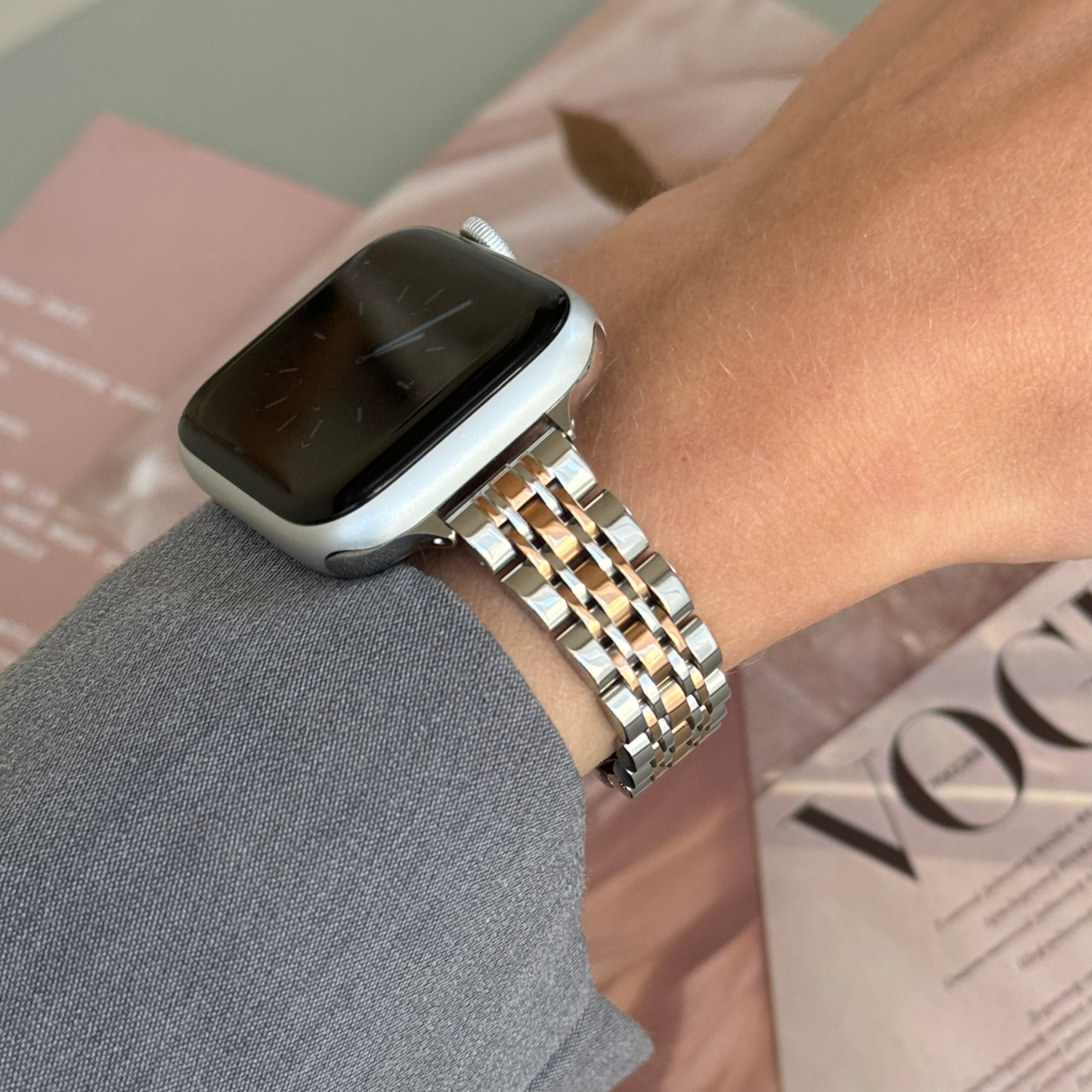Silver rose gold woven link Apple Watch band worn with a grey sleeve beside a fashion magazine, stainless steel bracelet for Apple Watch 38mm to 49mm