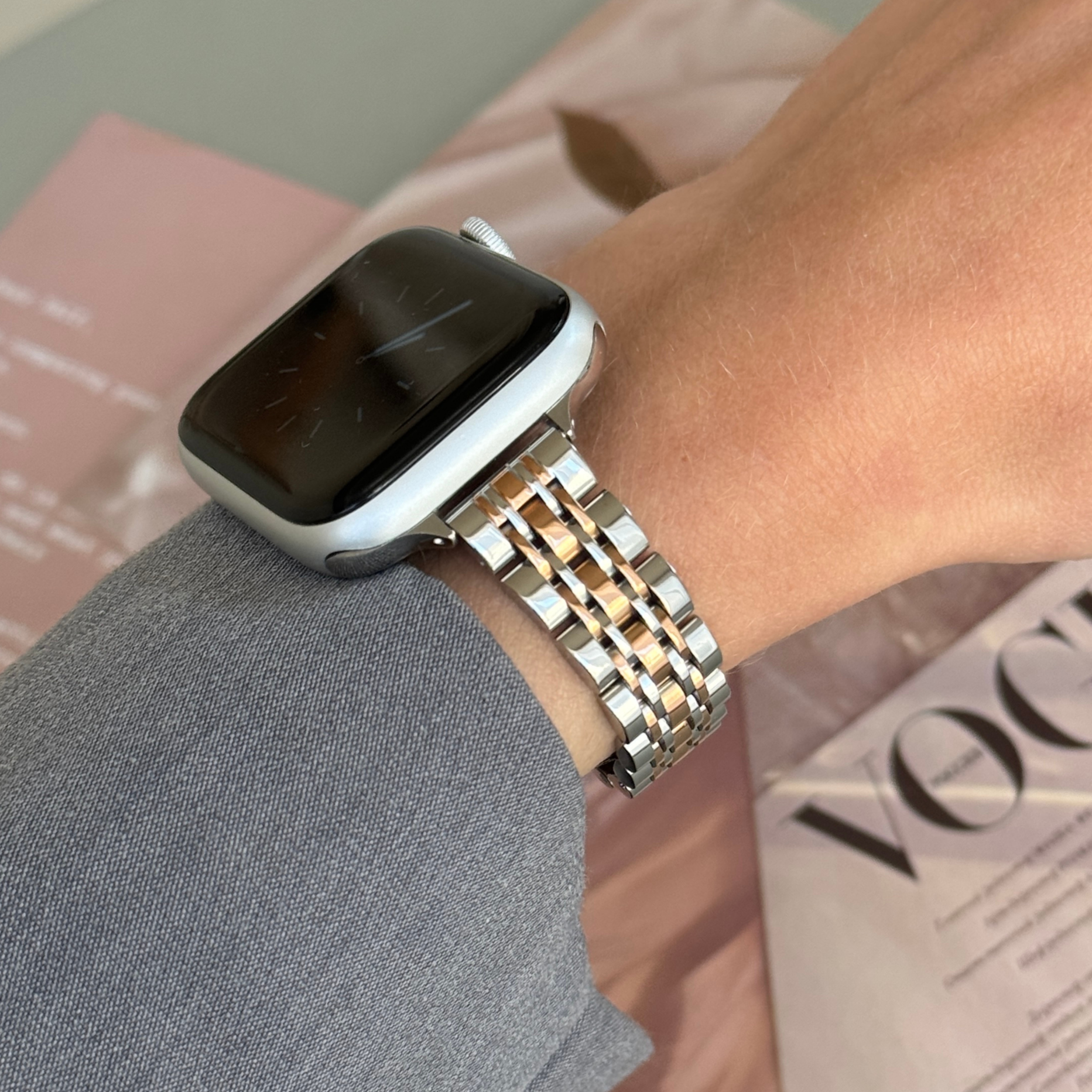 Silver rose gold woven link Apple Watch band worn with a grey sleeve beside a fashion magazine, stainless steel bracelet for Apple Watch 38mm to 49mm