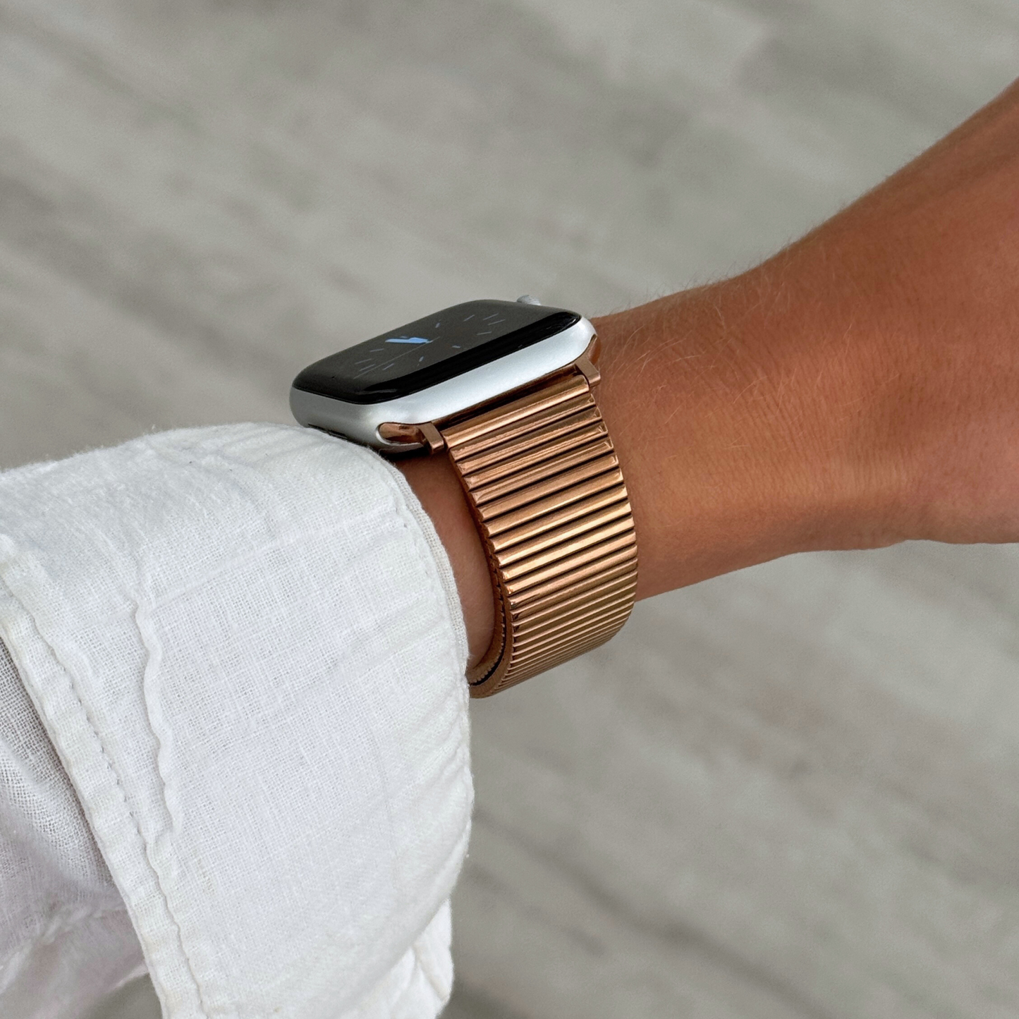 Rose gold ribbed loop Apple Watch band worn on a wrist with a white linen sleeve. Chic stainless steel iWatch strap for a perfect fit, compatible with Apple Watch 38mm to 49mm, Series 1-11, SE and Ultra