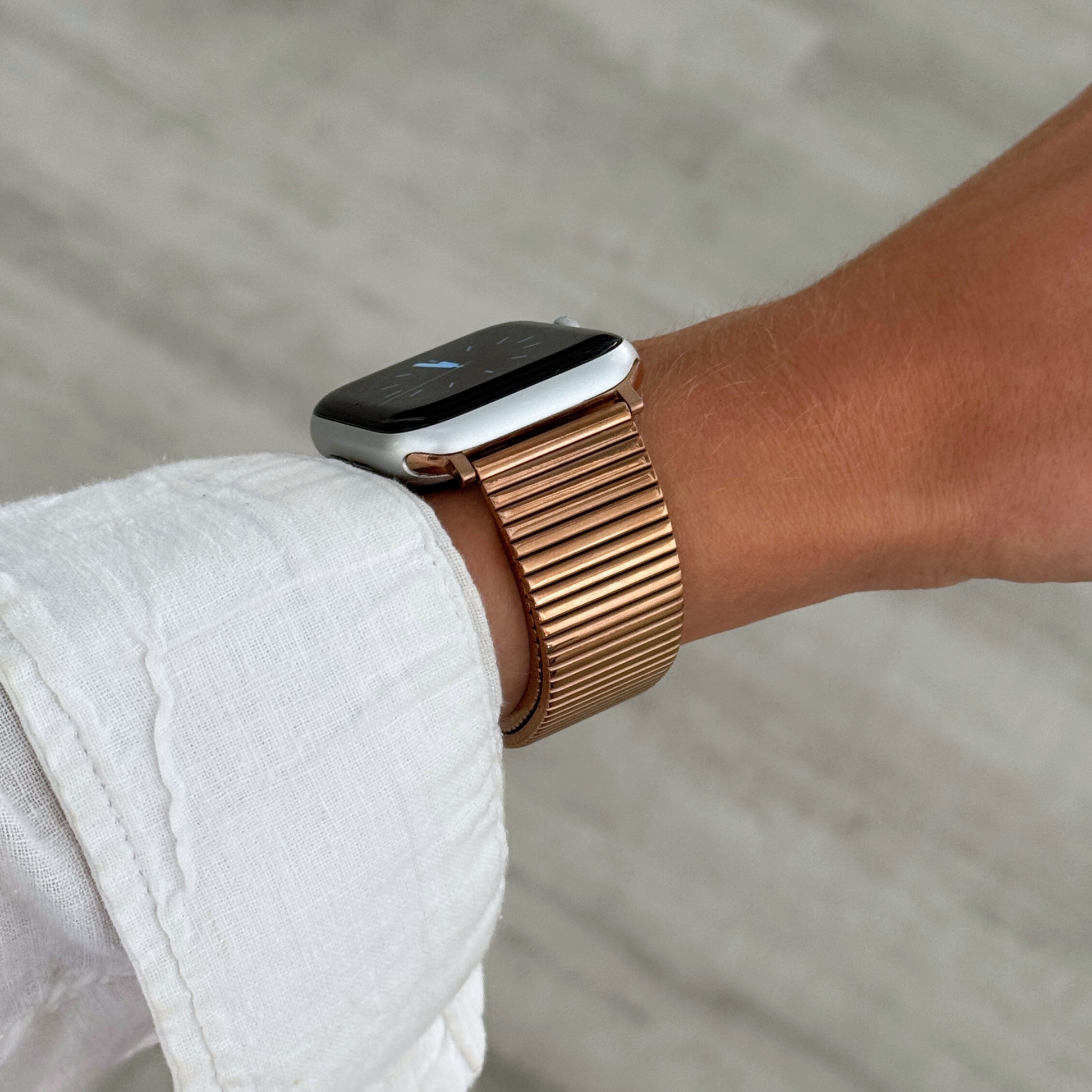 Rose gold ribbed loop Apple Watch band worn on a wrist with a white linen sleeve. Chic stainless steel iWatch strap for a perfect fit, compatible with Apple Watch 38mm to 49mm, Series 1-11, SE and Ultra