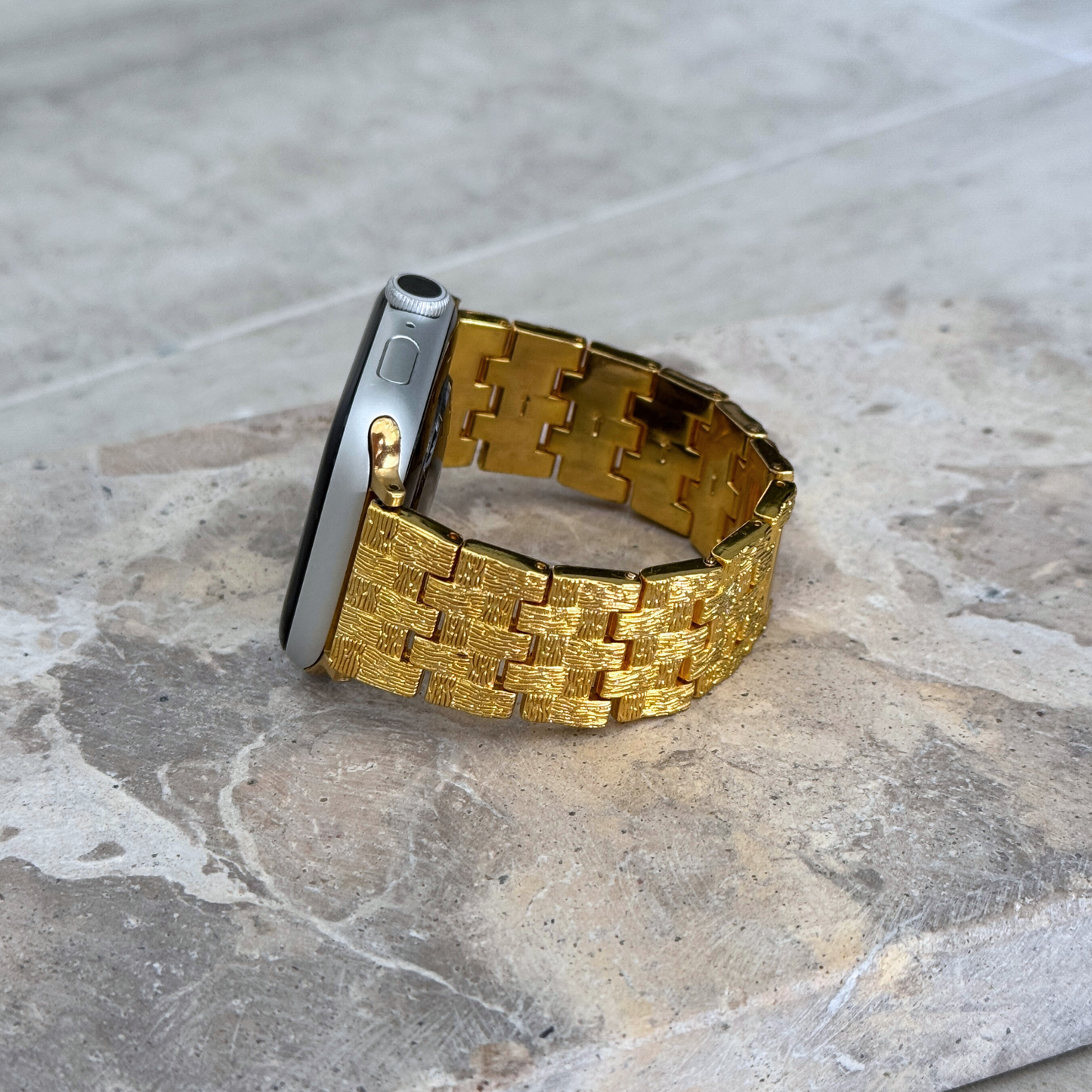Gold baroque pattern Apple Watch band propped upright on a stone surface. Metal iWatch strap with brushed matte and polished shine, compatible with Apple Watch 38mm to 49mm, Series 1-11, SE and Ultra