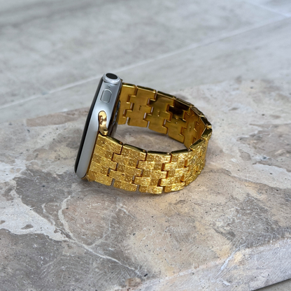 Gold baroque pattern Apple Watch band propped upright on a stone surface. Metal iWatch strap with brushed matte and polished shine, compatible with Apple Watch 38mm to 49mm, Series 1-11, SE and Ultra