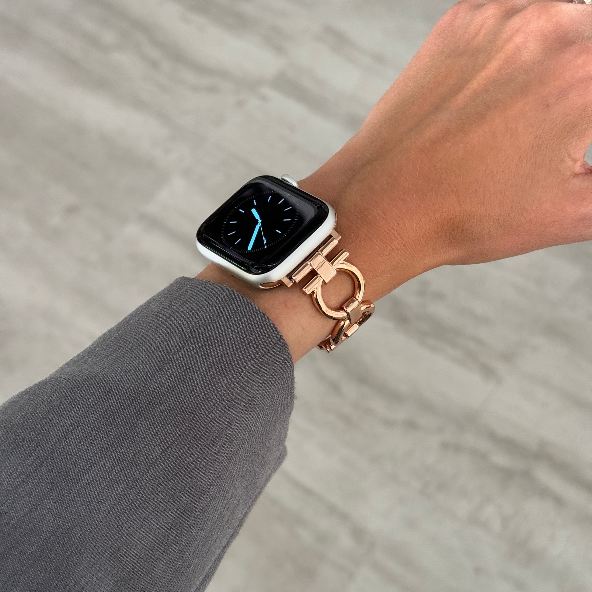 Rose gold circle link Apple Watch band worn on a wrist above grey trousers. Slim metal iWatch strap for everyday wear, compatible with Apple Watch 38mm to 49mm, Series 1-11, SE and Ultra